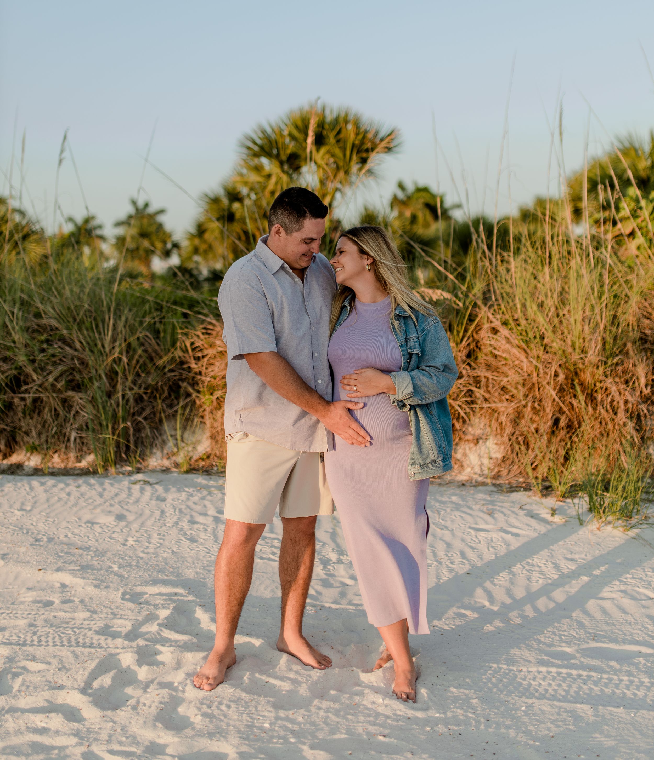 A couple stands together on sandy beach with tropical vegetation in the warm sunset light.