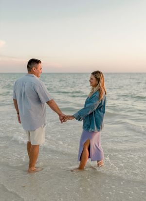 A couple holding hands while standing in gentle ocean waves during a sunset beach photoshoot.