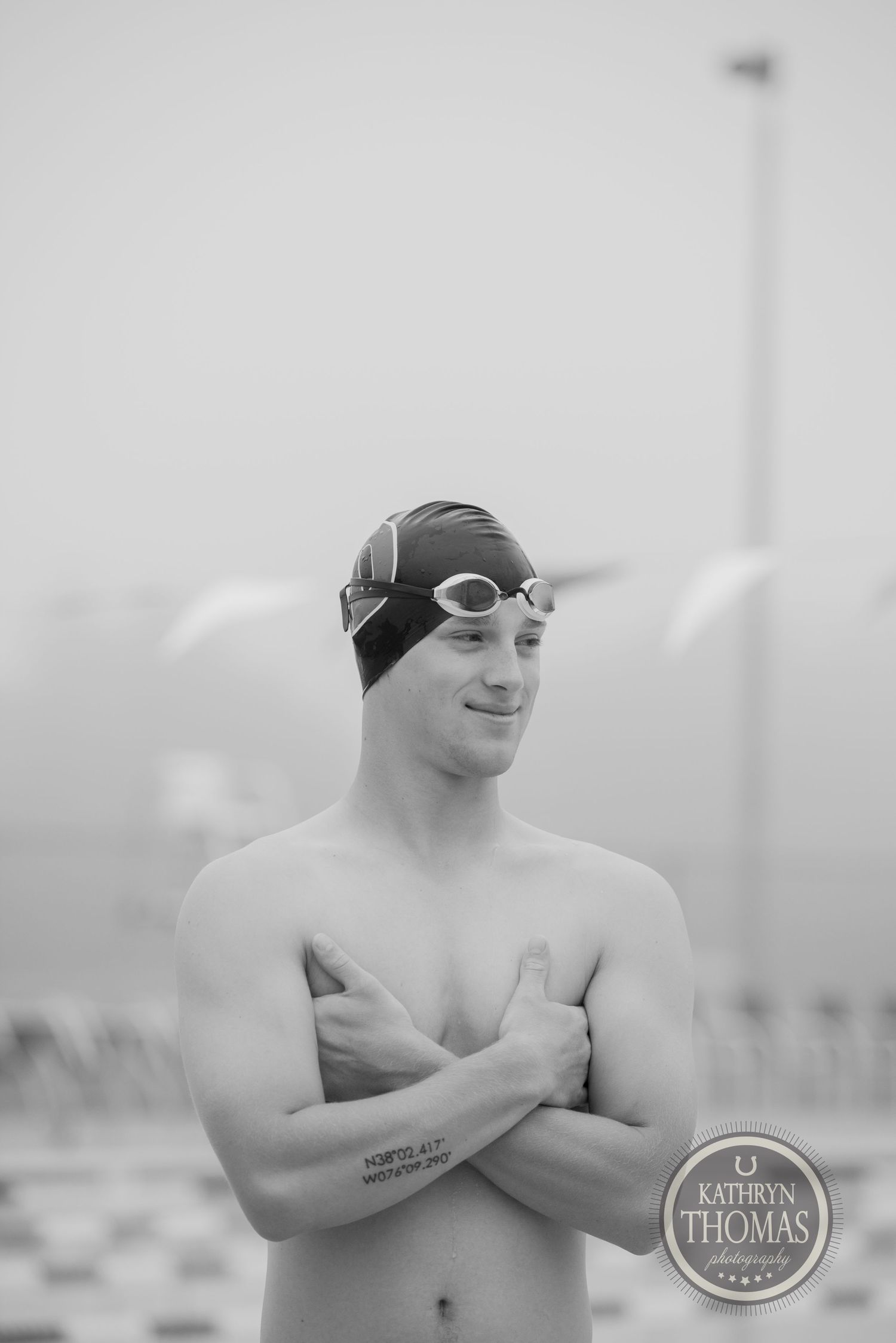 A competitive swimmer wearing a swim cap and goggles poses with arms crossed by the pool.