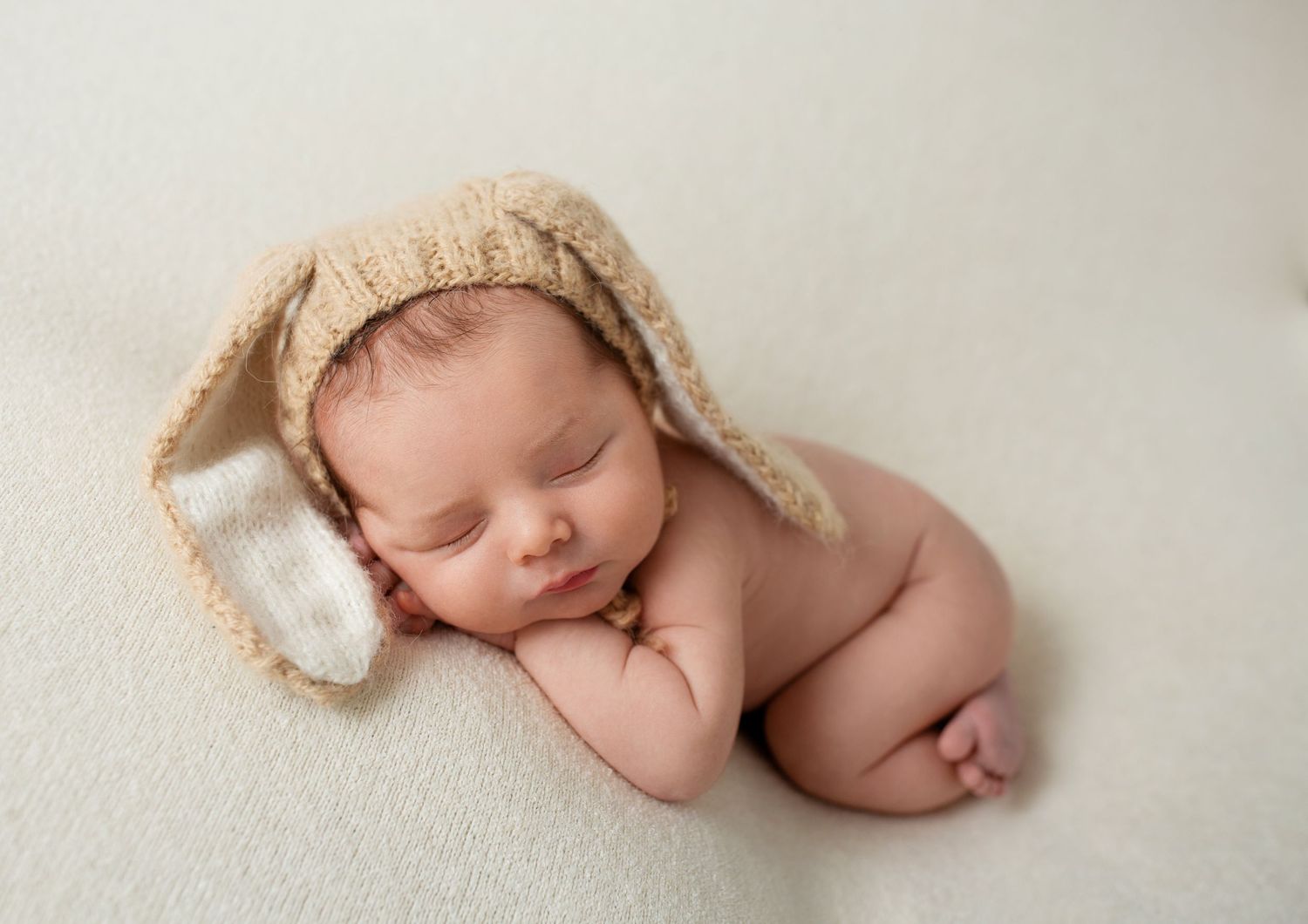 Sleeping newborn baby wearing knit bunny hat poses on cream colored backdrop.