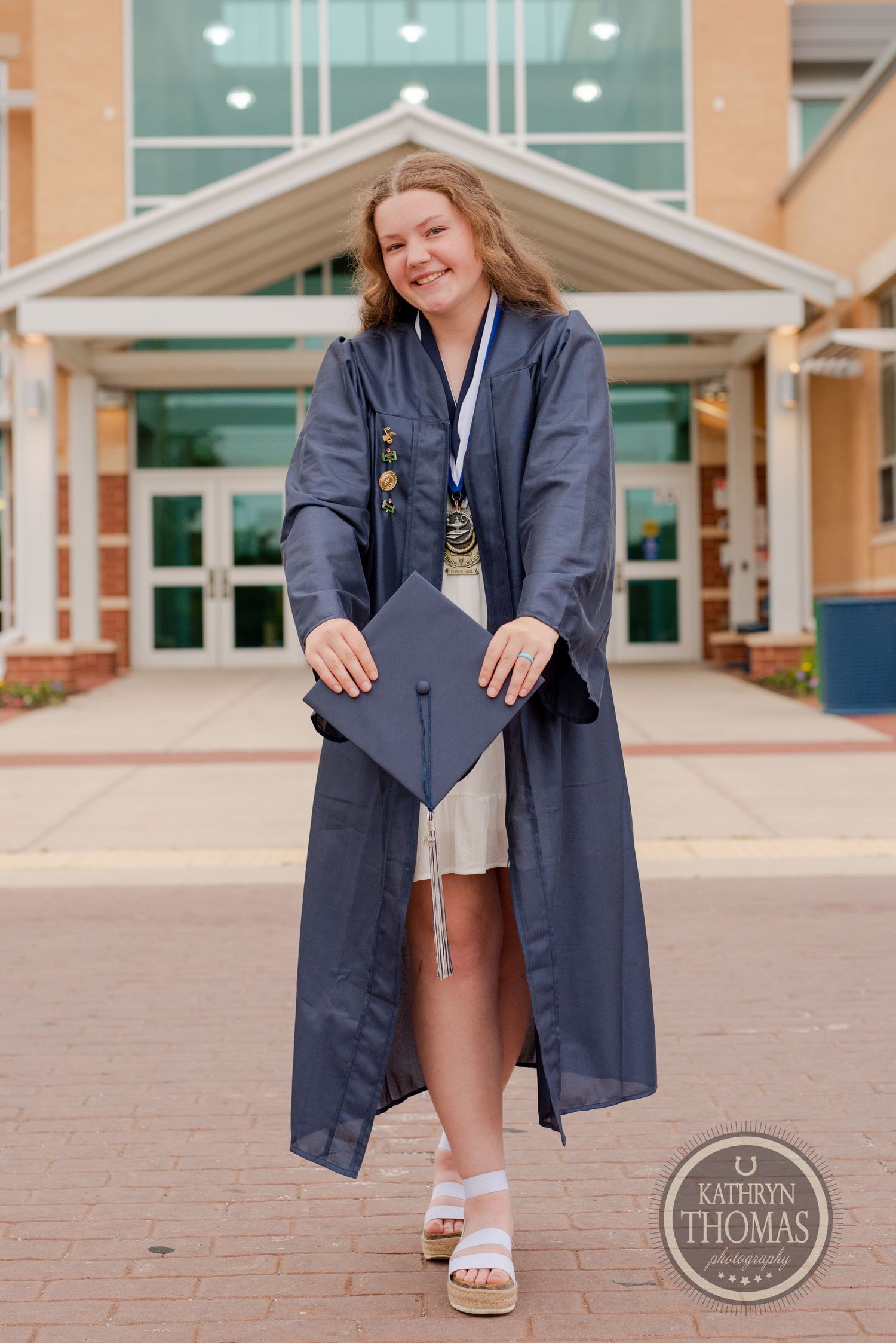 Graduate holds mortarboard cap while posing in front of school building entrance.