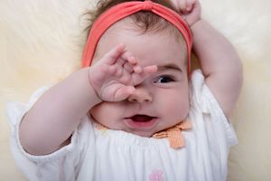 Baby wearing white outfit and coral headband playfully covers face with hand while smiling.