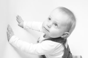Black and white portrait of baby in white clothing leaning against wall.