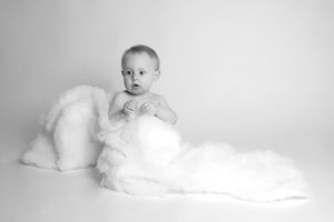 Artistic black and white portrait of infant sitting on soft white blanket.
