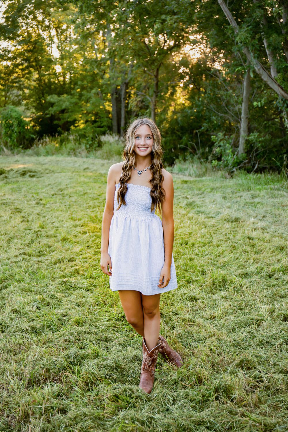 A summer portrait in a white sundress and brown boots standing in a grassy field at golden hour.