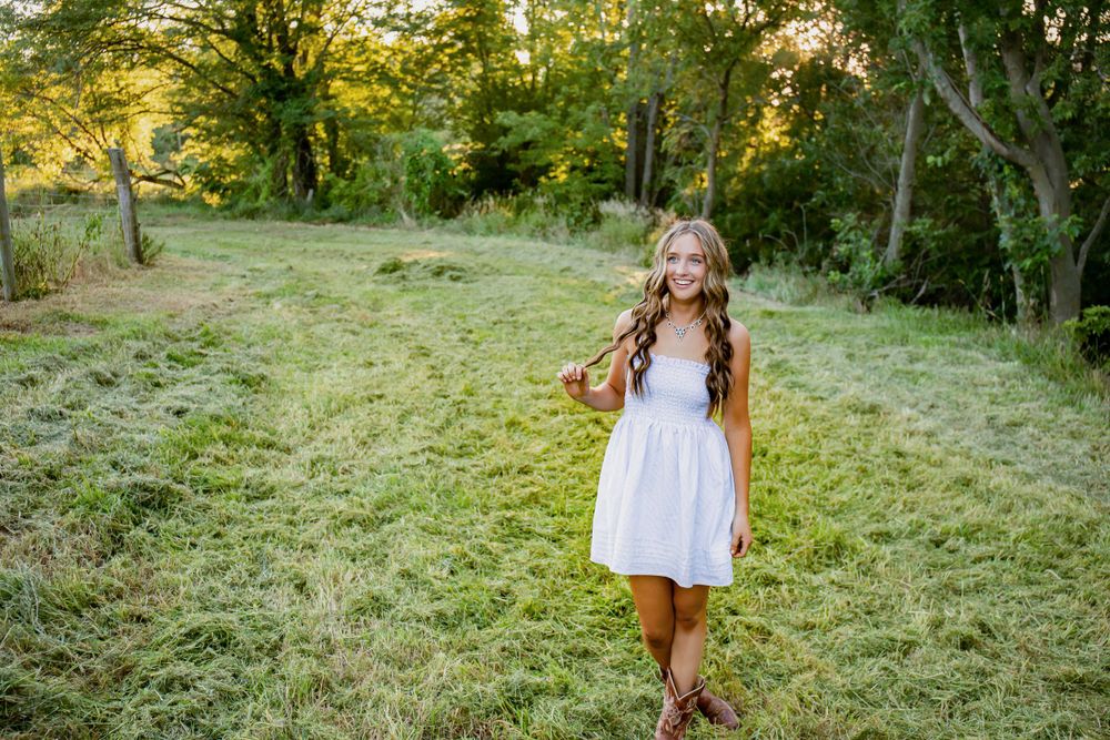 Natural light portrait of someone twirling in a white dress among green grass and trees.