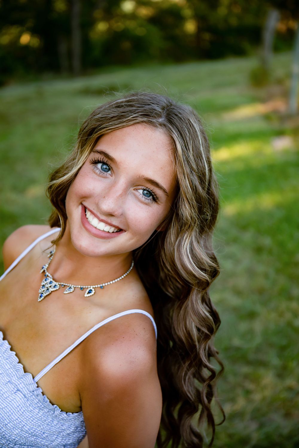 Portrait of someone wearing a white dress and crystal necklace against a sunlit green background.