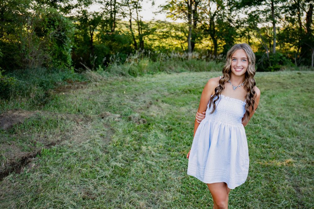 Figure in a white dress poses against lush greenery and trees at dusk.