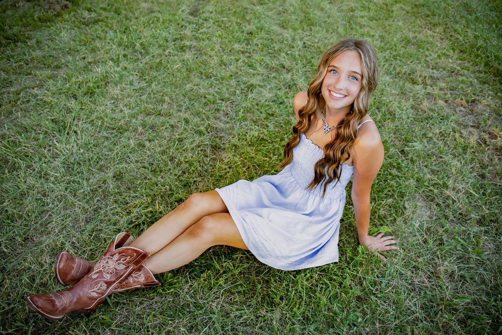Casual pose in white dress and brown boots on grassy ground.