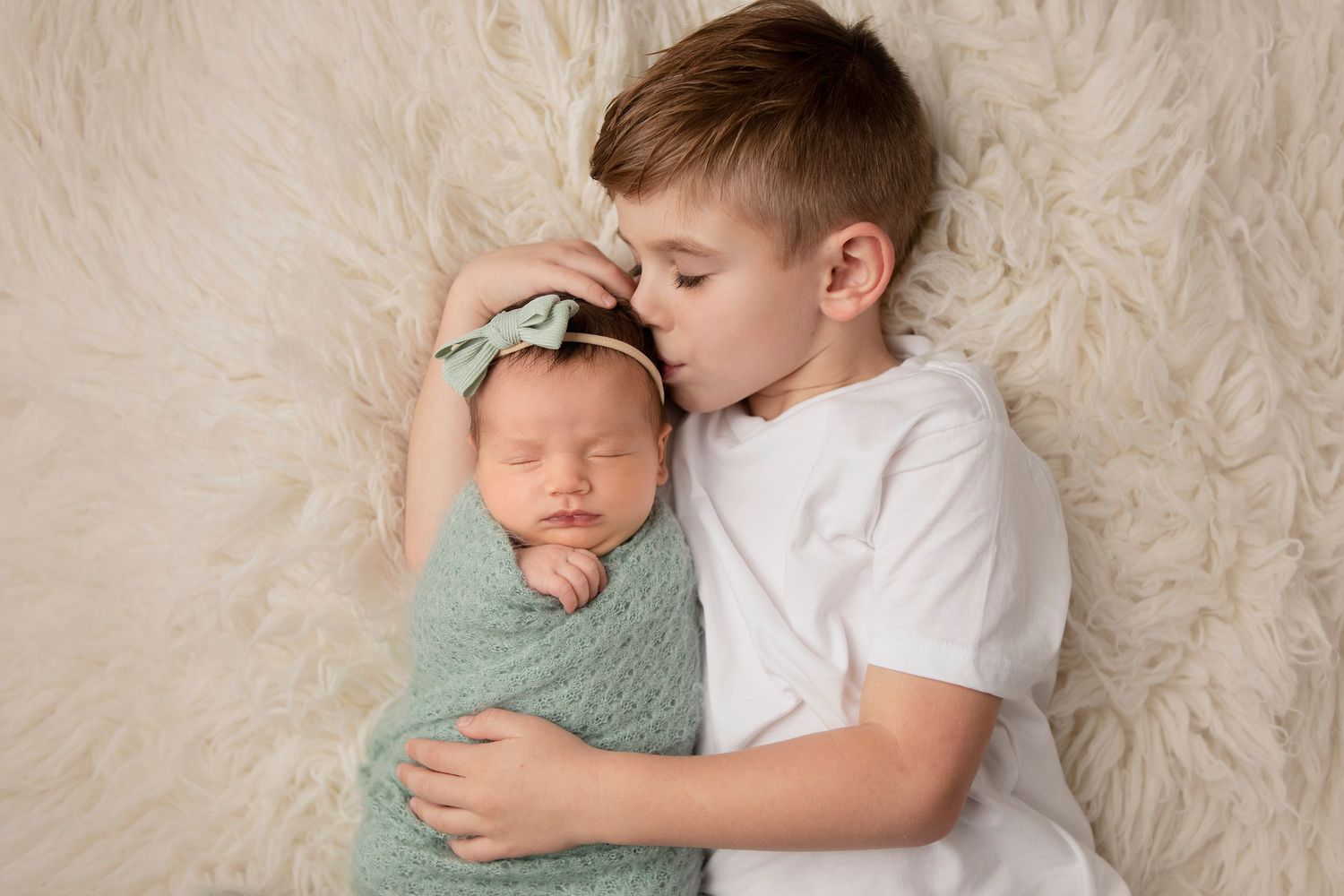 Newborn sibling portrait on cream fur backdrop showing tender moment.