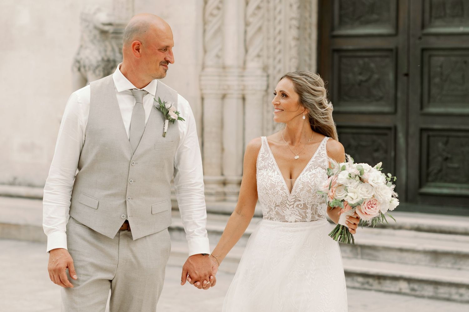 Elegant couple in light-colored wedding attire walking hand in hand by ornate architecture.