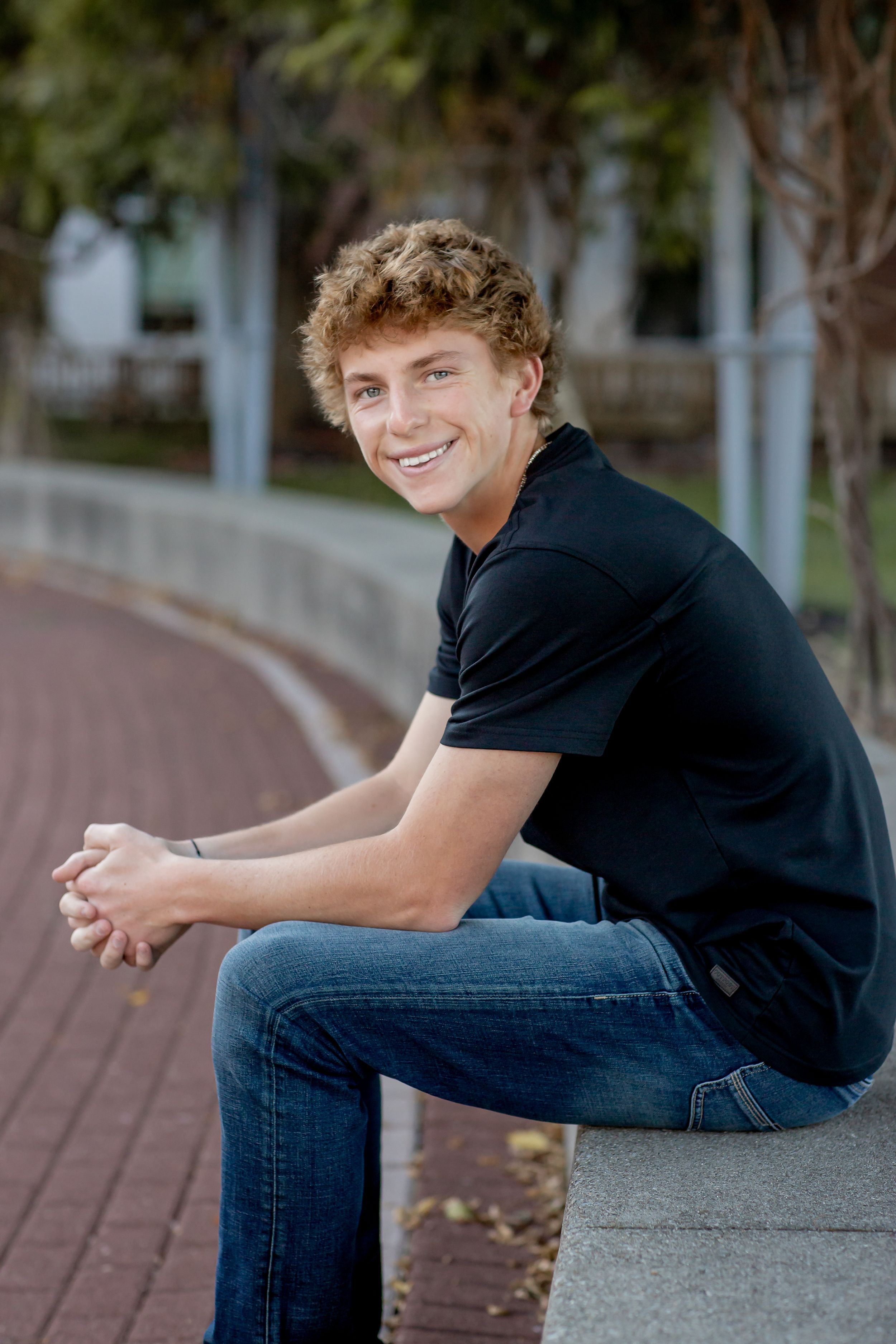 Smiling teenage boy in a black polo shirt sits on a ledge along a tree-lined walkway.