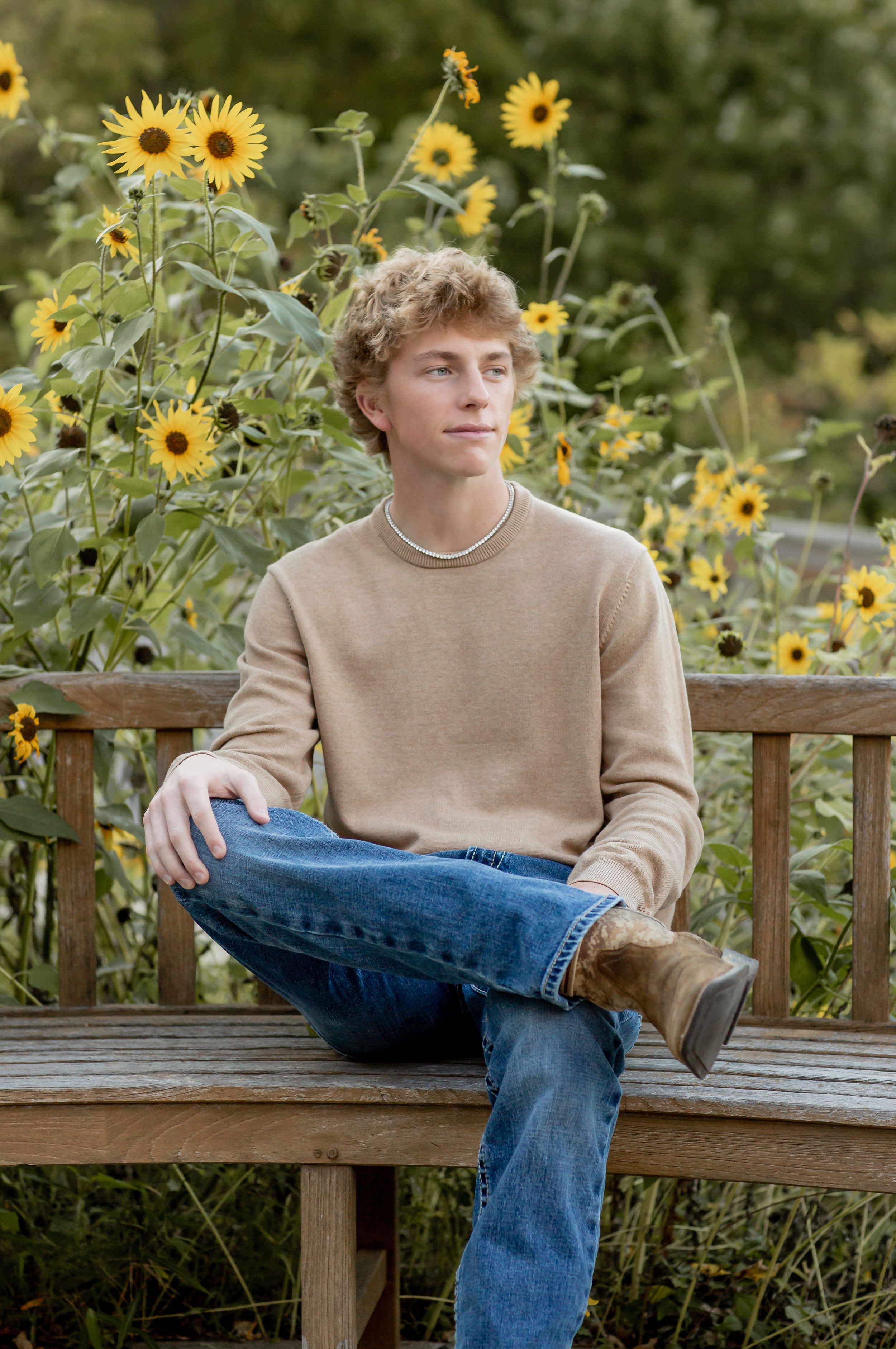 Teen boy with curly blonde hair sitting on a wooden bench surrounded by bright yellow sunflowers outdoors.