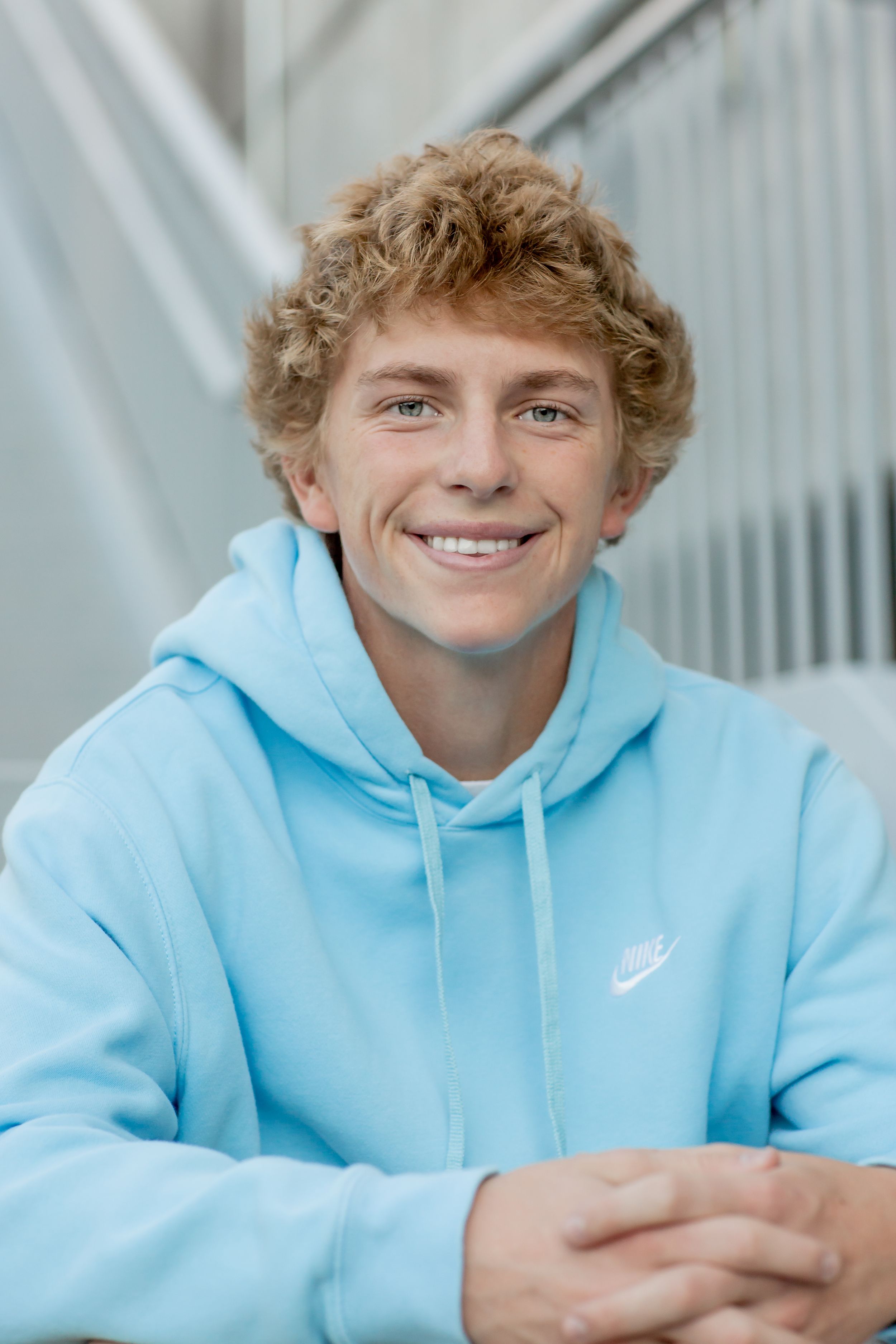 Smiling teenage boy with curly blond hair wears a light blue Nike hoodie indoors.