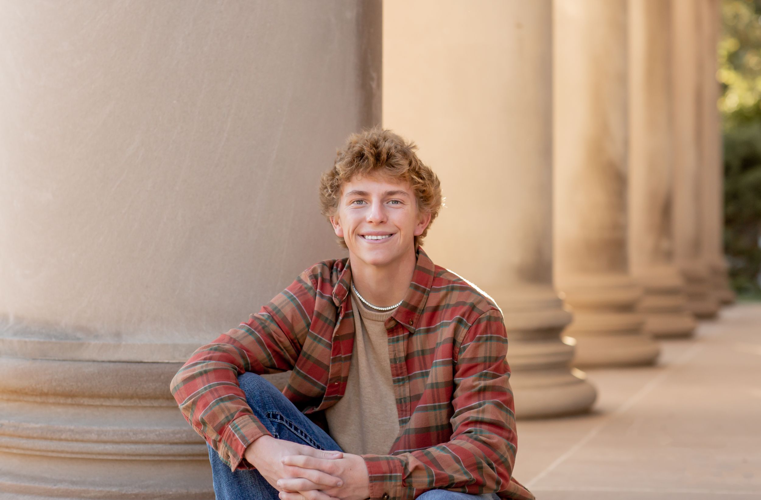 Smiling teenage boy in a plaid shirt sits on steps beside large stone columns outdoors.