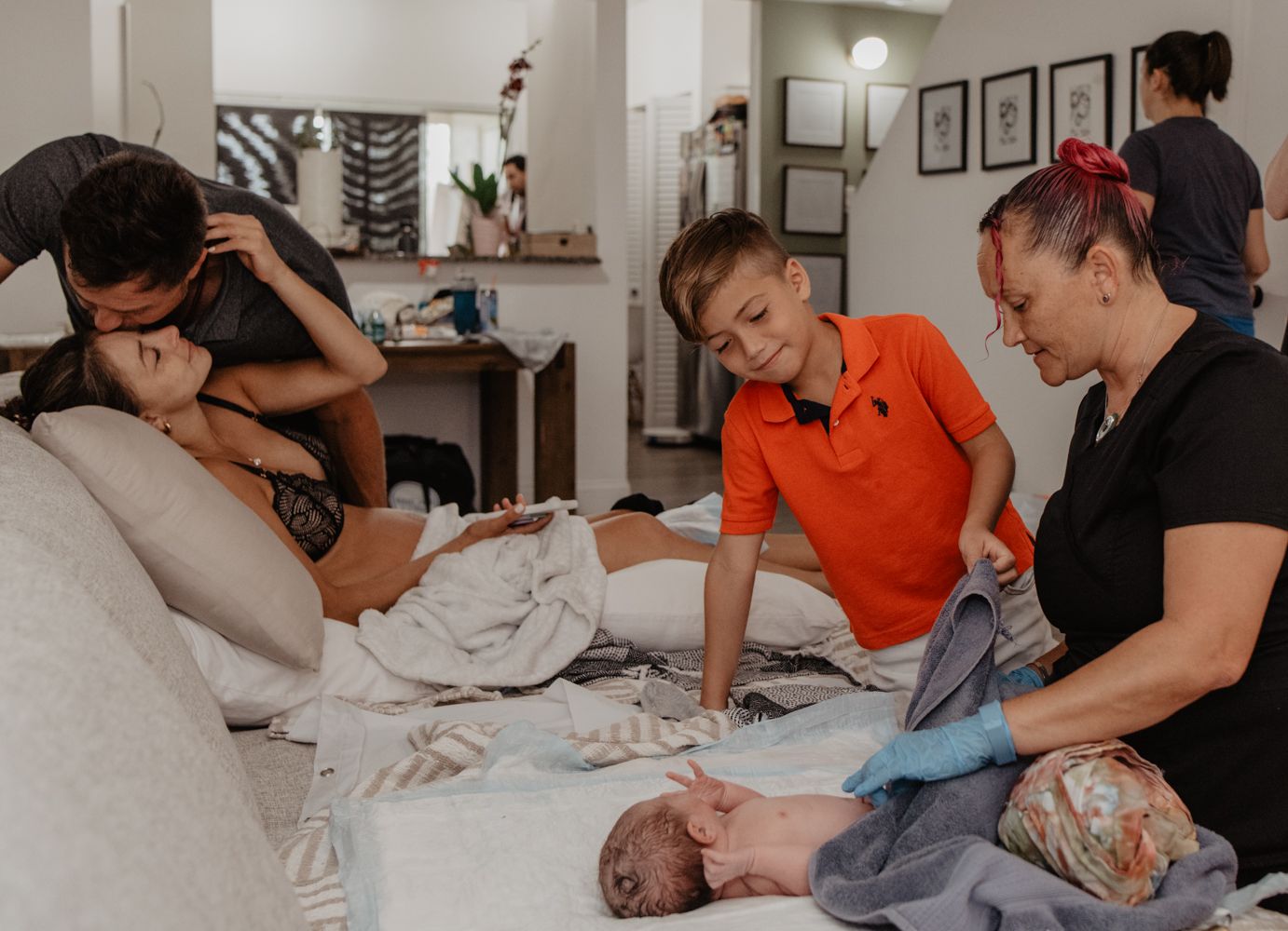 family, post birth, sitting on the couch. dad kissing mom's forehead, while little boy and midwife look at the baby