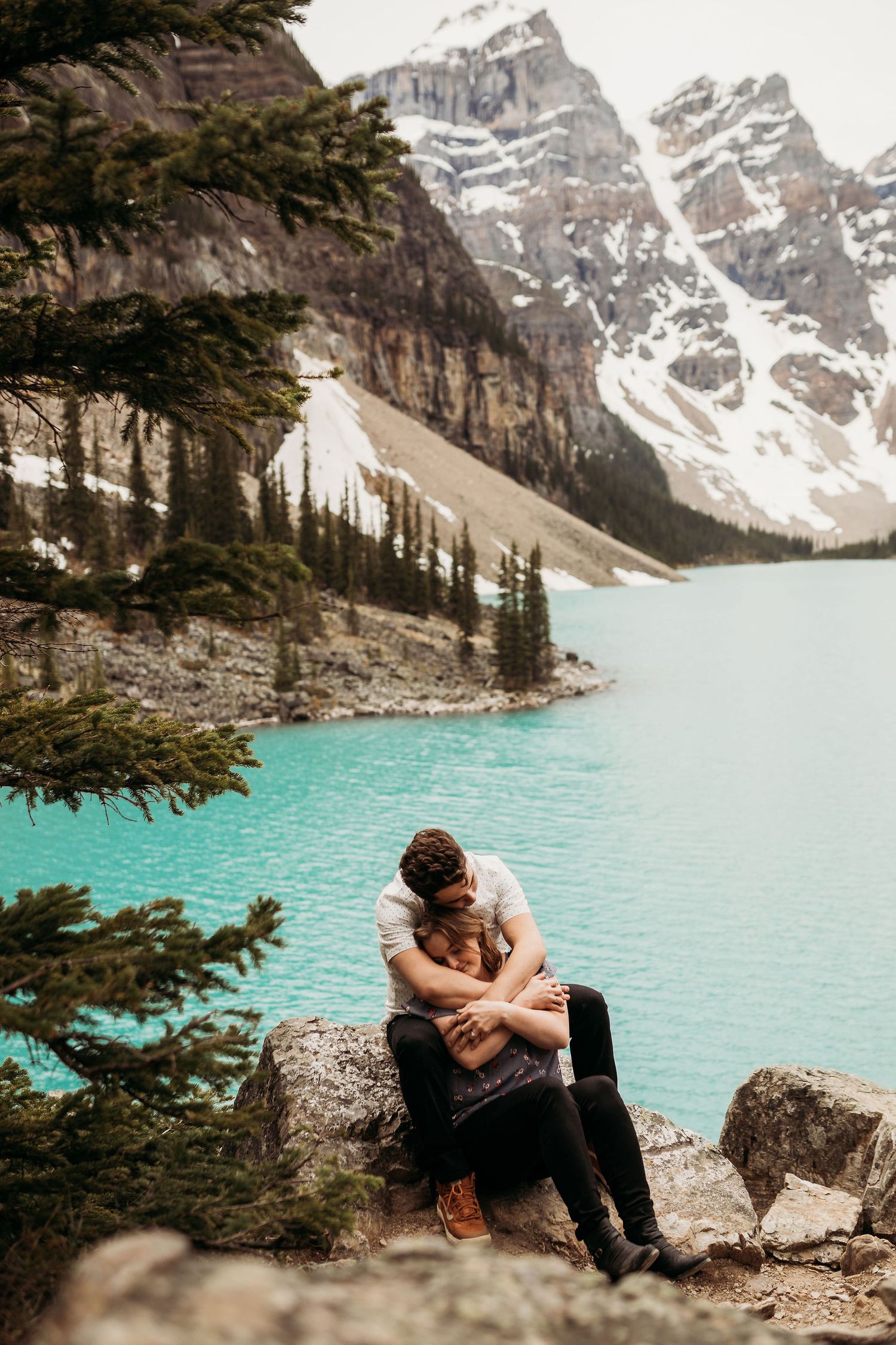 A couple sits on rocks overlooking the pristine turquoise waters of Moraine Lake in Banff.
