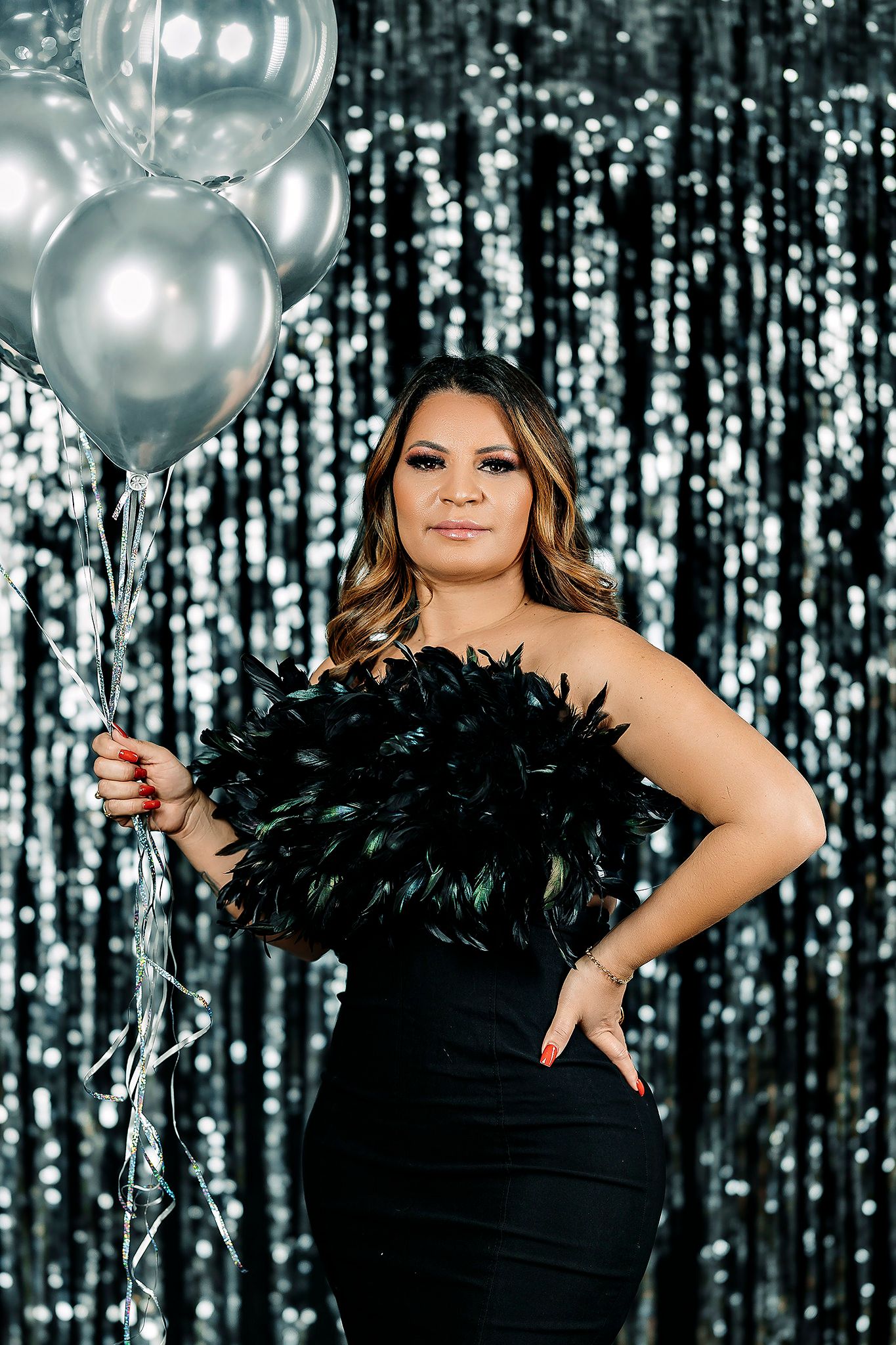 Festive portrait with black feather dress and silver balloons against metallic curtain.