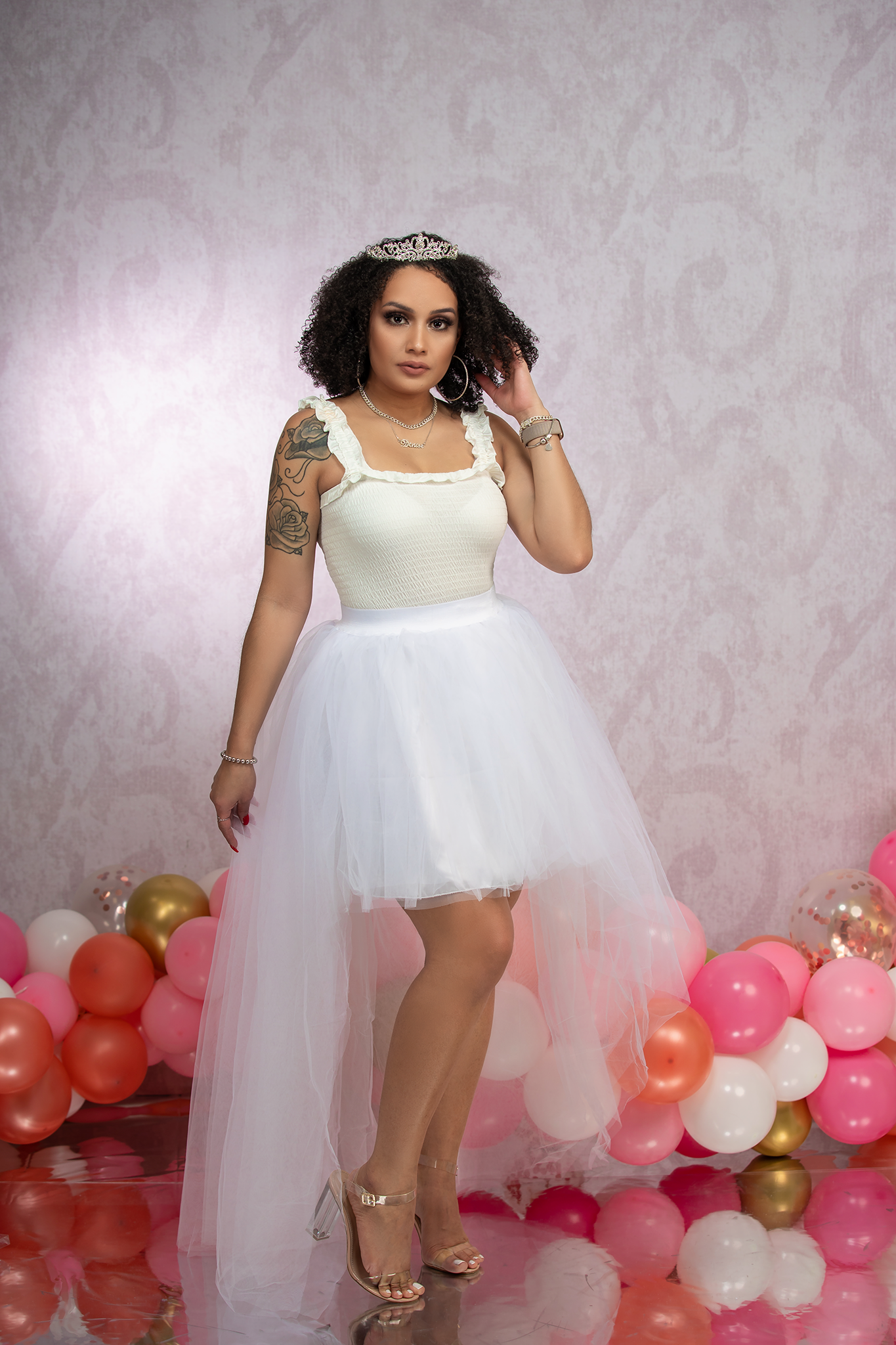 Person in white high-low tulle dress poses with tiara against pink balloon backdrop.