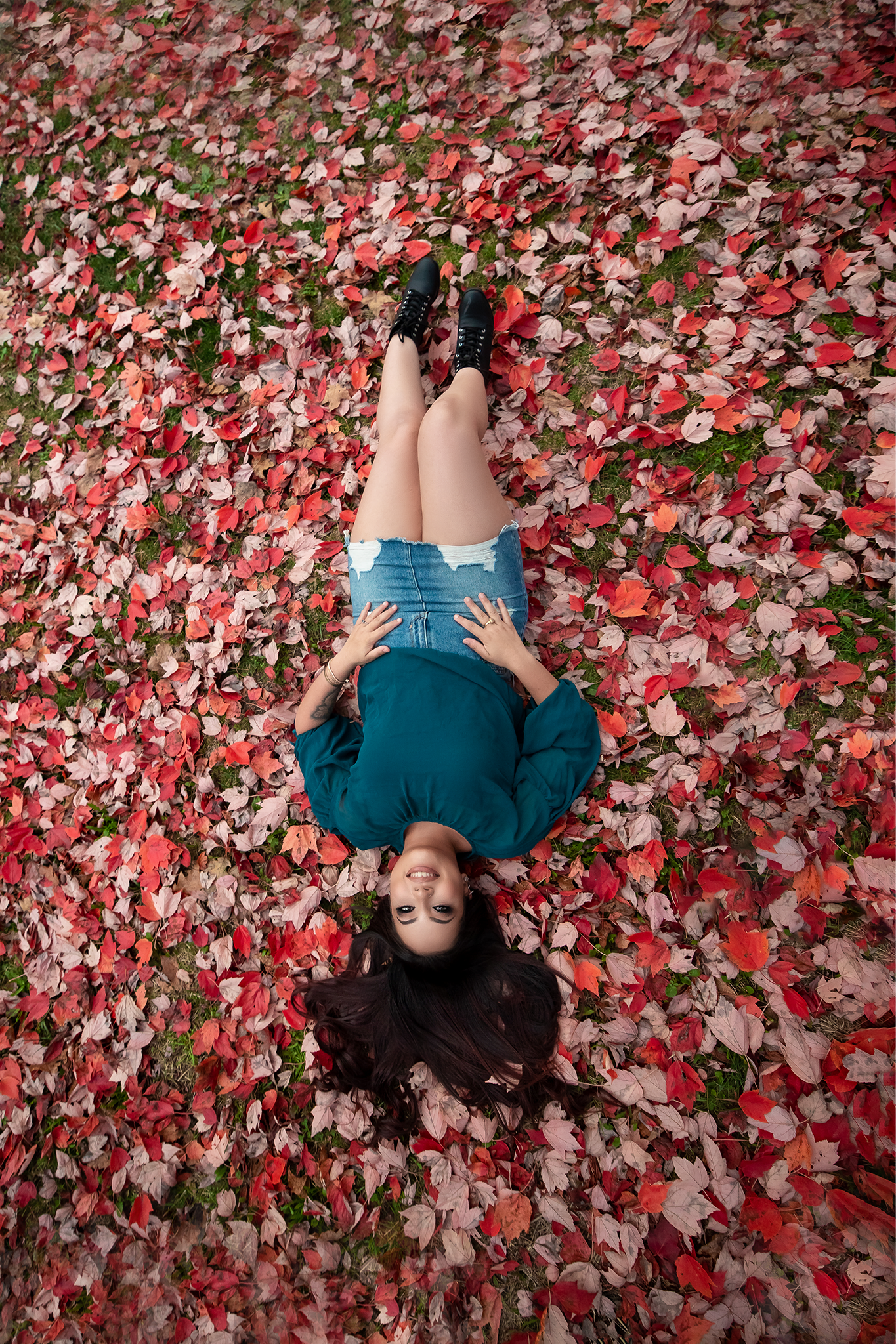 Person lying on a bed of bright red autumn leaves in a teal outfit.