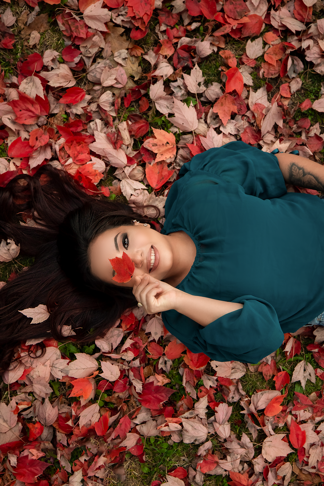 Person in teal dress poses on red autumn leaves during fall photo session.