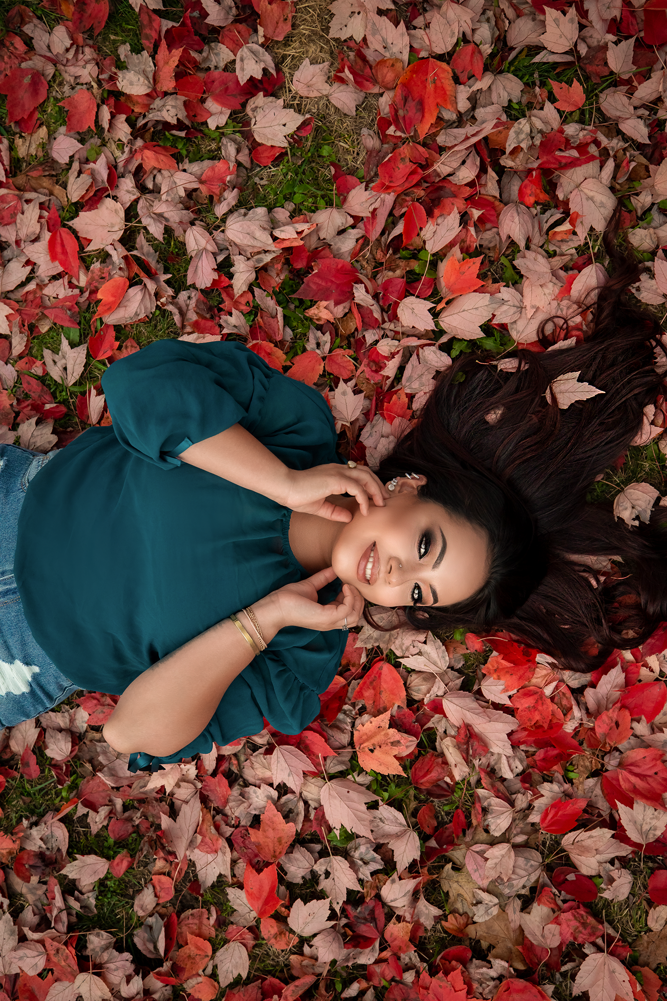 Person in dark teal dress lies surrounded by red and orange fall leaves.