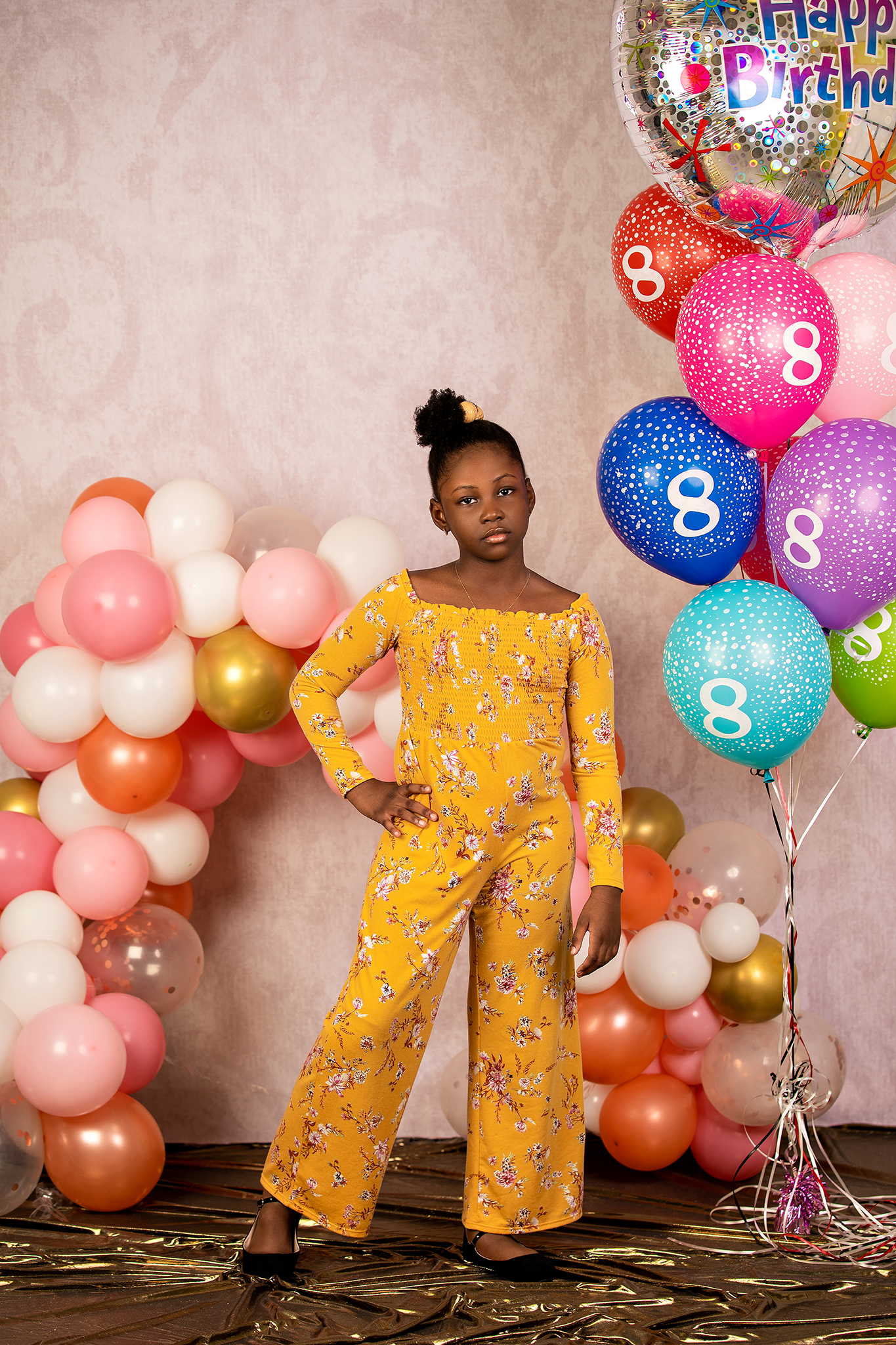 Young person in yellow floral jumpsuit poses with birthday balloons showing number 8.