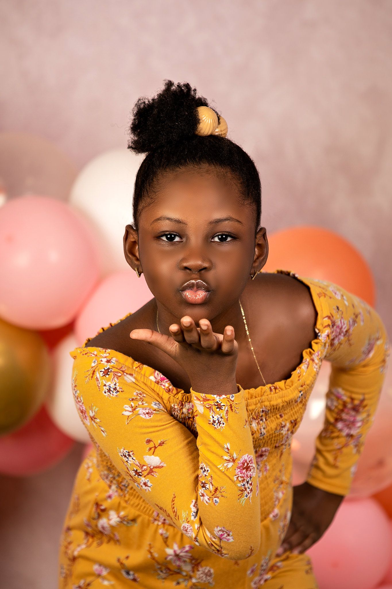 Child in yellow floral dress poses against pink backdrop with balloons.