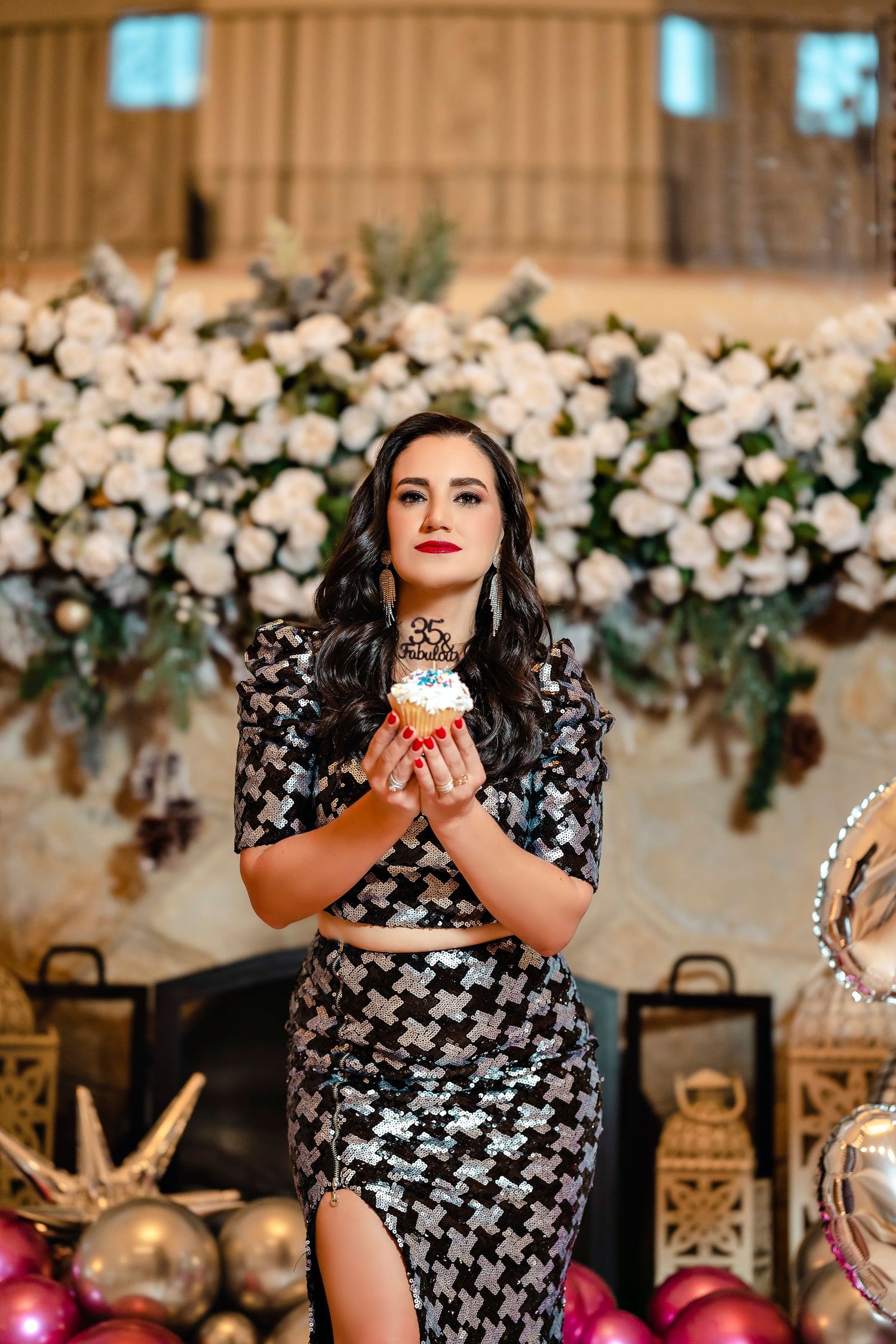 Woman in patterned dress holds cupcake against floral backdrop at celebration event.