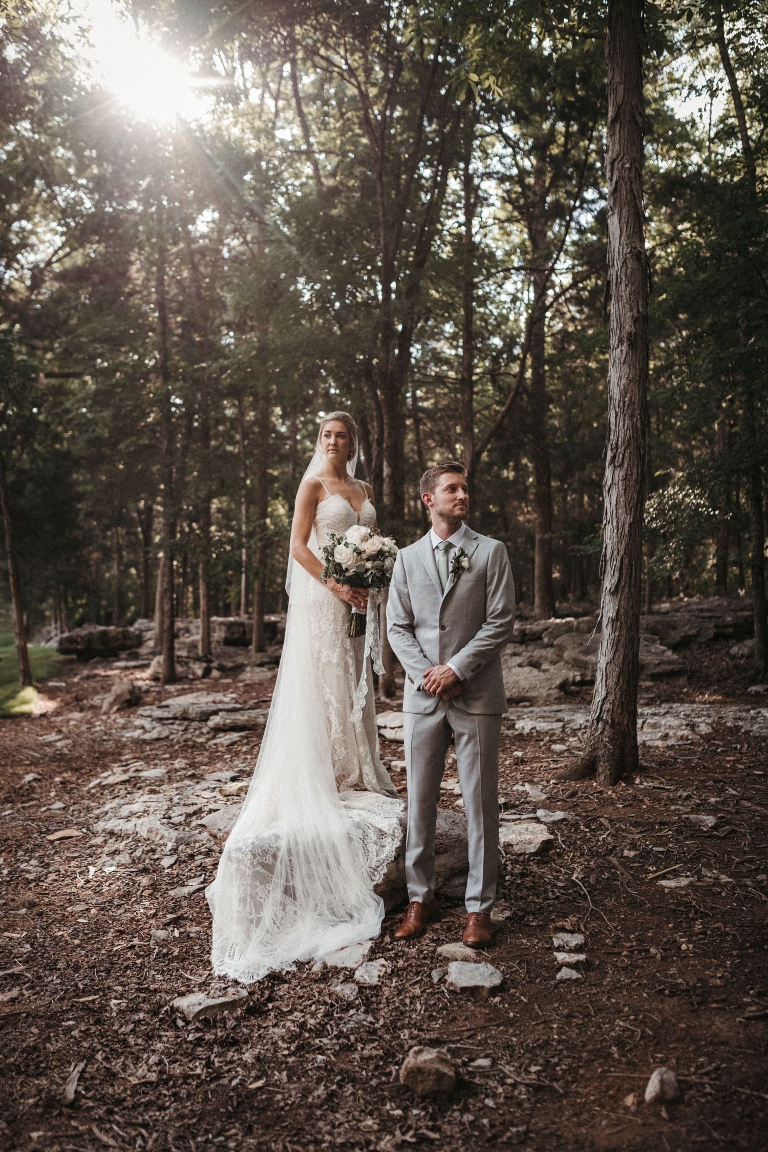 Wedding couple poses in sunlit wooded area wearing grey suit and lace gown.