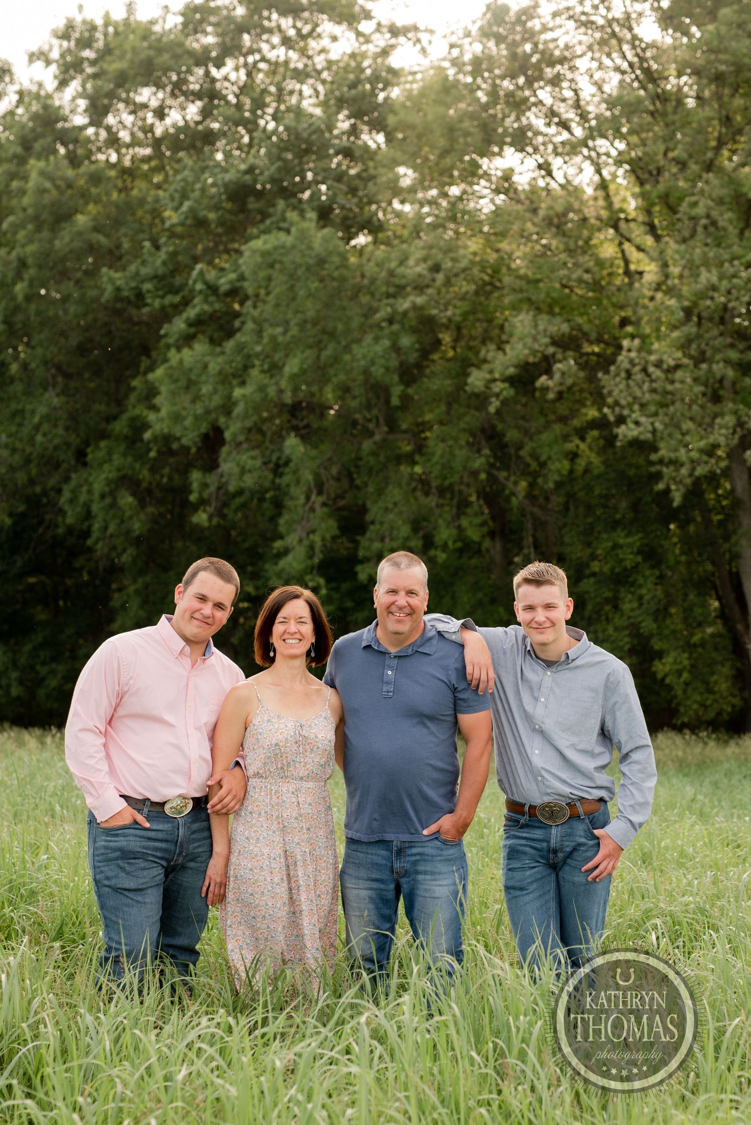 Family portrait in casual attire standing together in tall grass with trees in background.