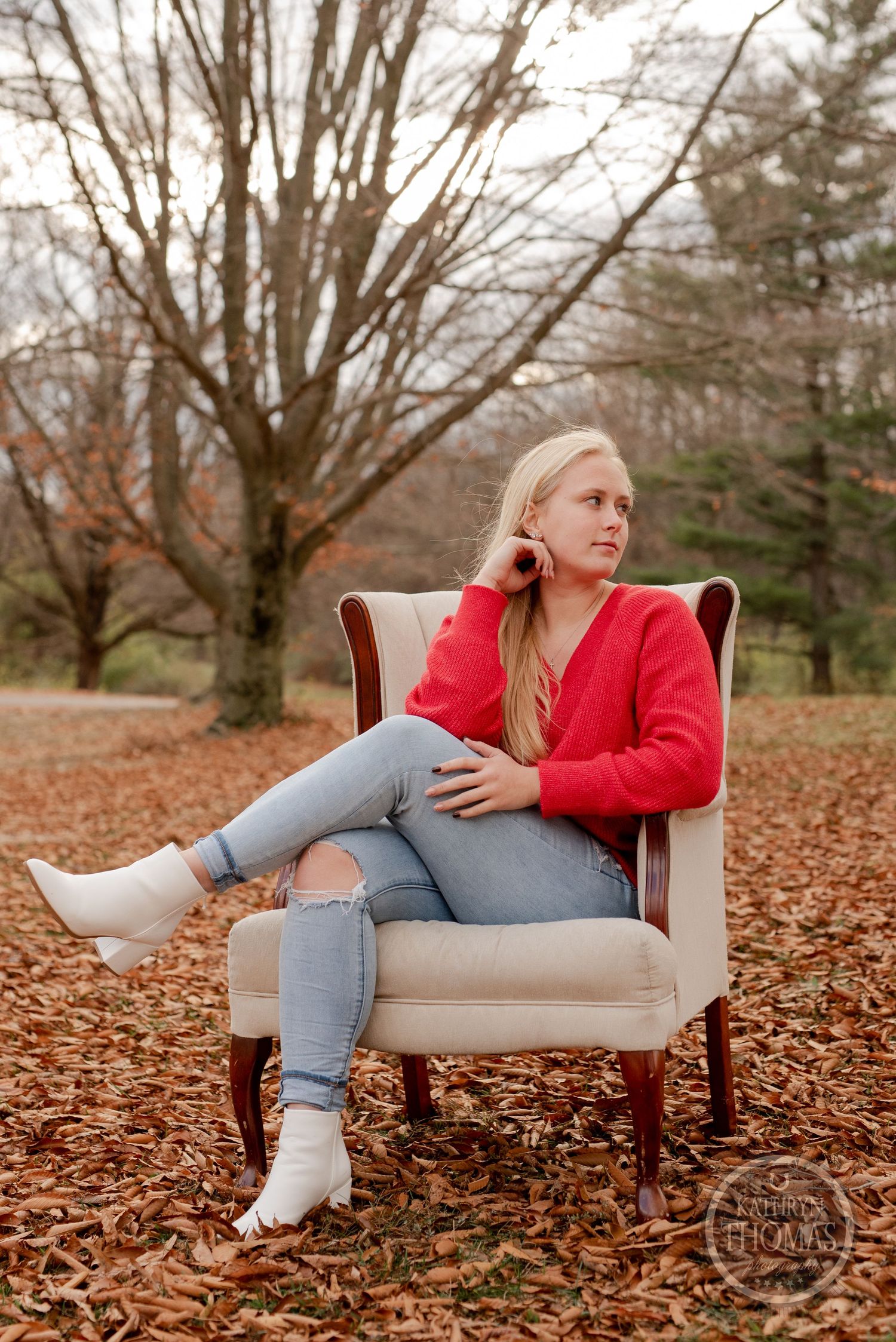 Relaxed pose in beige armchair wearing red sweater and distressed jeans among fall foliage.