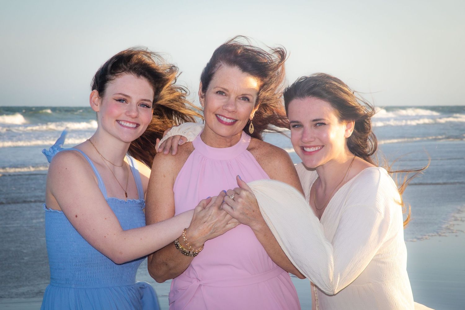 Three women in pastel dresses embrace on a sunny beach.