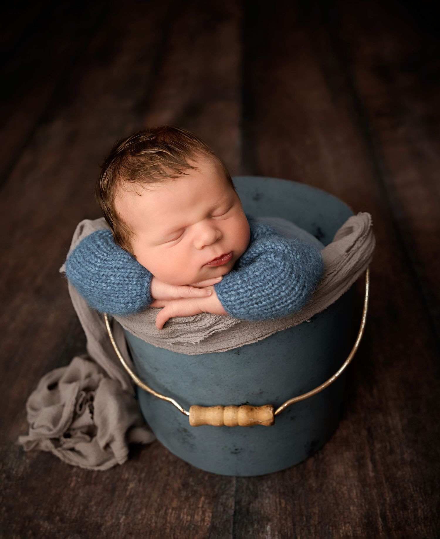 A peaceful sleeping newborn baby boy in a blue knit wrap sits posed inside a rustic teal bucket.