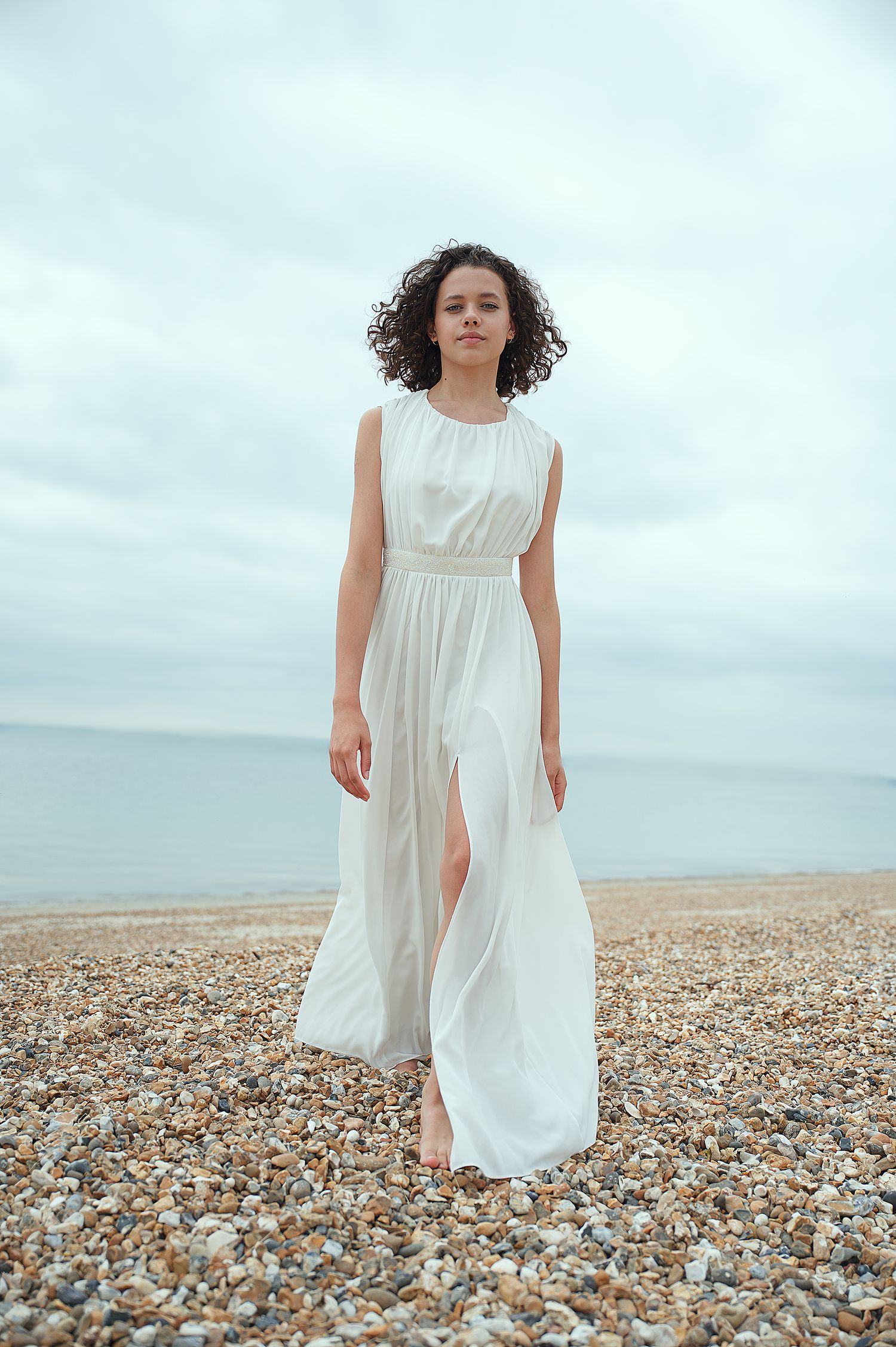 Model in elegant white maxi dress poses on seaside beach with natural curls and overcast sky.