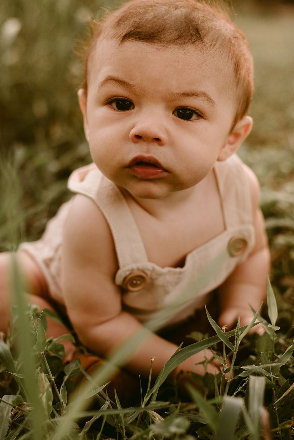 Close-up portrait of baby in beige overalls sitting among green foliage.