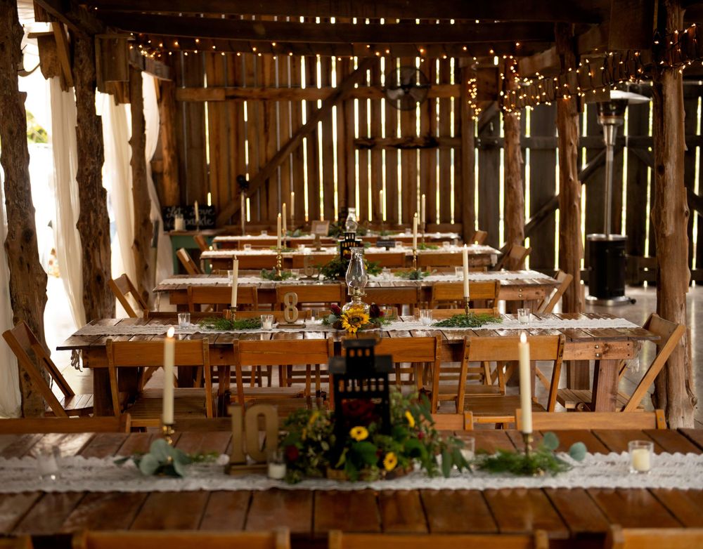 Long wooden tables set for a rustic wedding reception inside a barn with string lights and floral centerpieces.