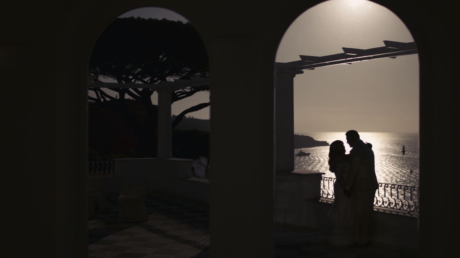 Silhouetted couple in an arched doorway against a sunset ocean view.