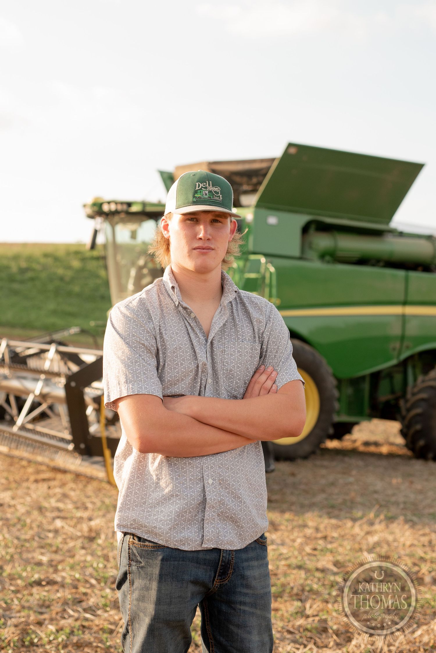 Farmer in baseball cap stands confidently in front of green harvesting equipment at sunset.