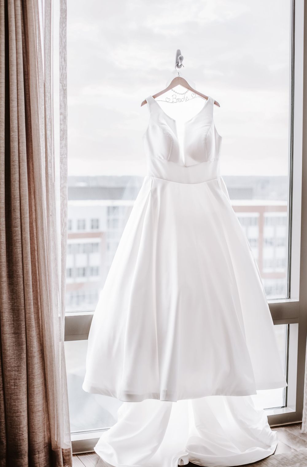 Multiple elegant white wedding dresses hanging in front of a bright window with brown curtains.