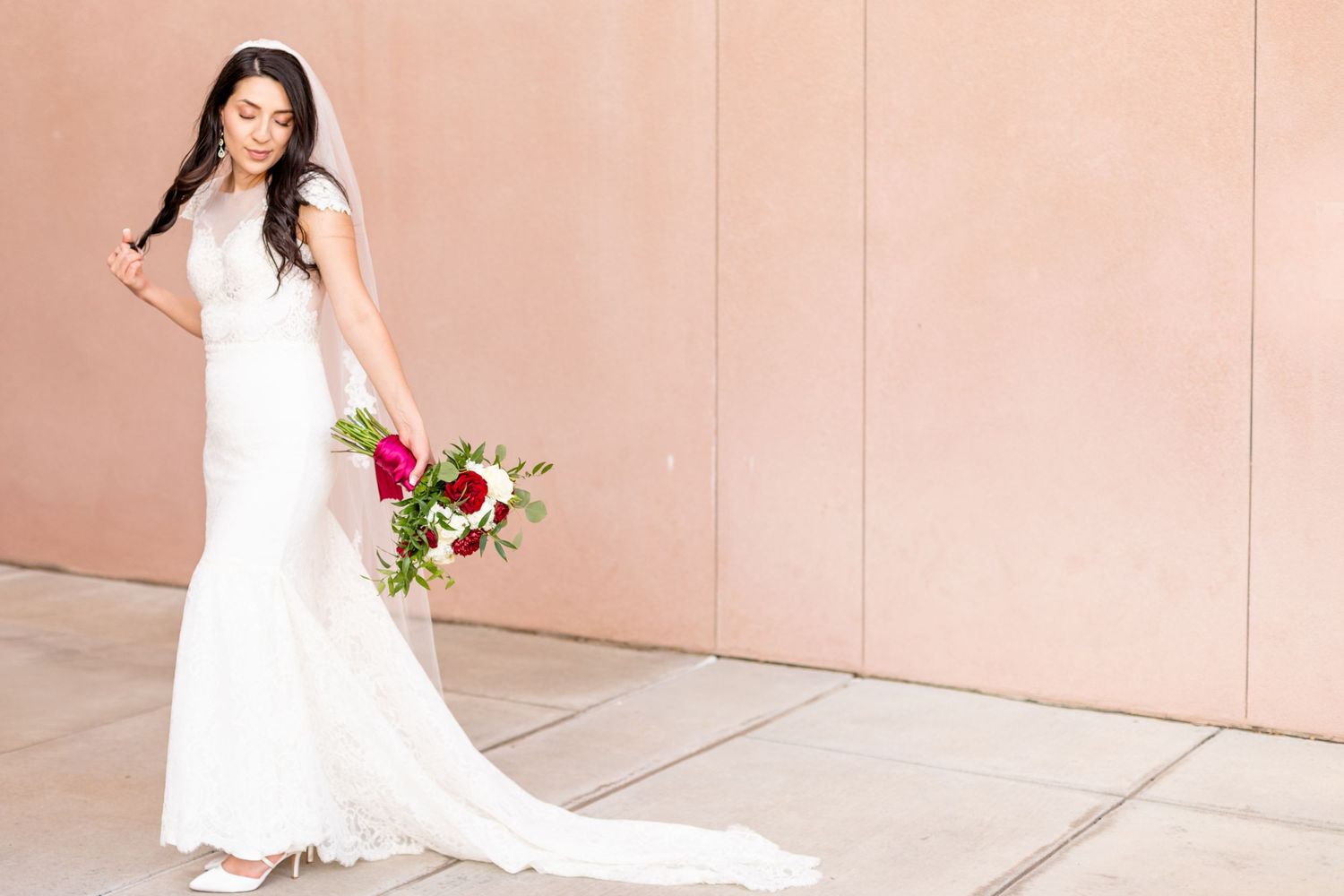 Bride in flowing lace gown poses with bouquet against peach colored wall.