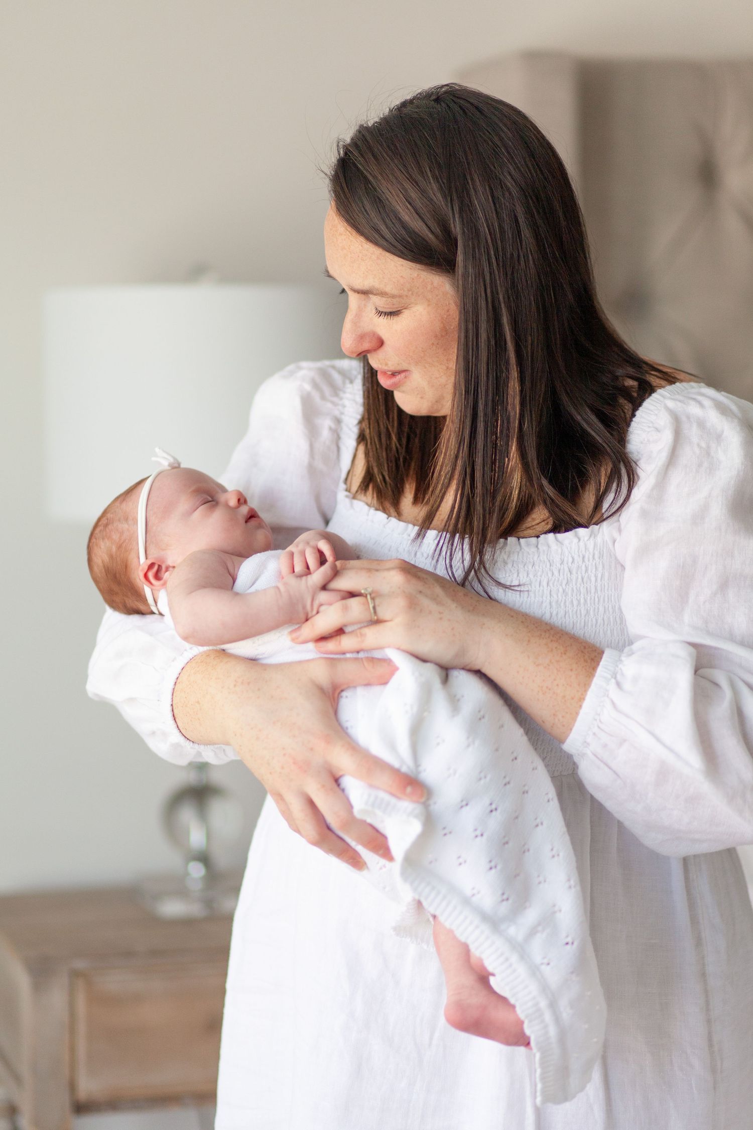 A brunette mother in a white dress holds her swaddled baby in her  Raleigh, NC in home newborn session.