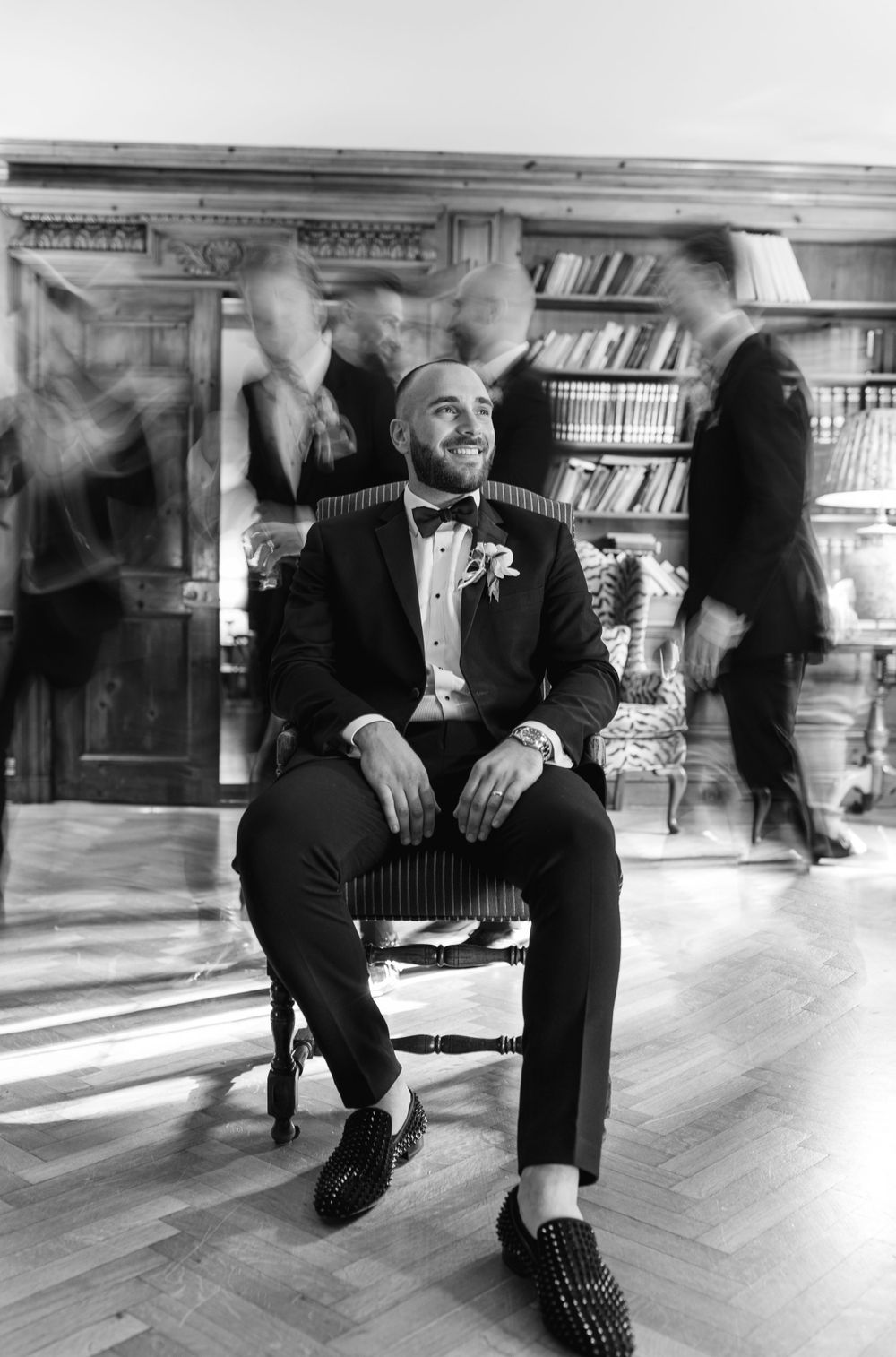A well-dressed man in a tuxedo sits casually in a library with motion-blurred figures in the background.