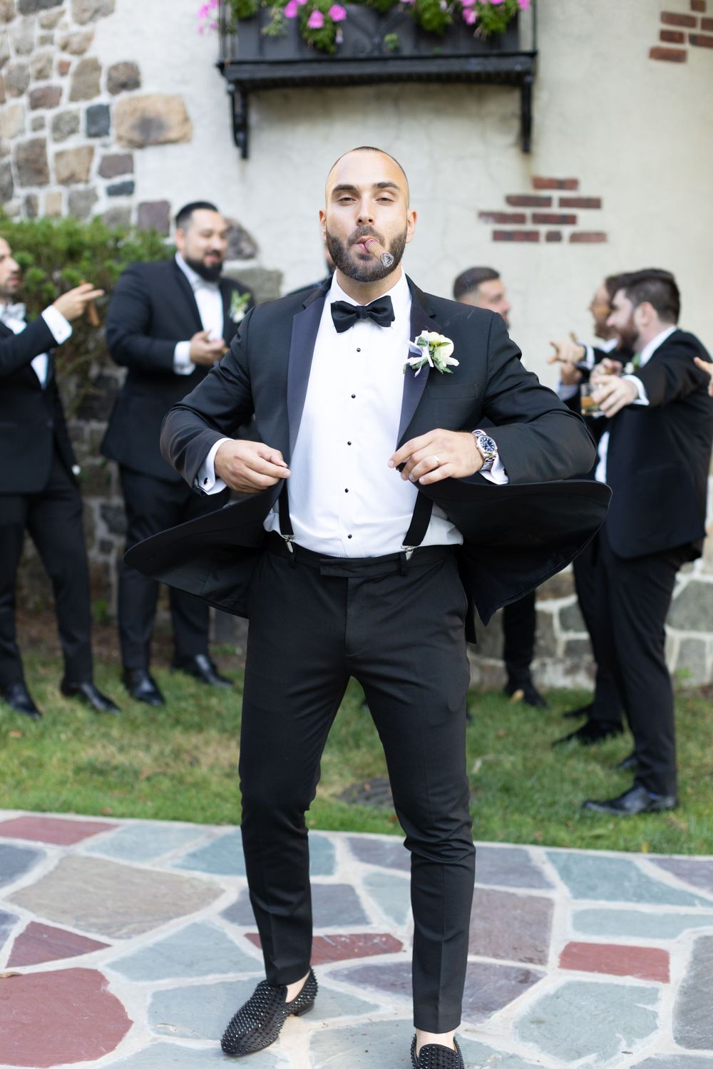 Groom in fitted black tuxedo poses confidently outside stone building.