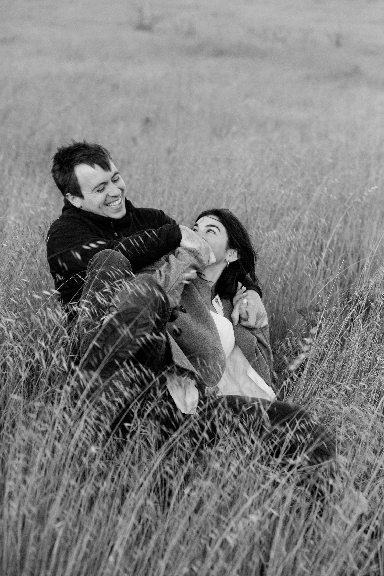 A couple shares a joyful, candid moment lying together in a field of tall grass, captured in black and white.