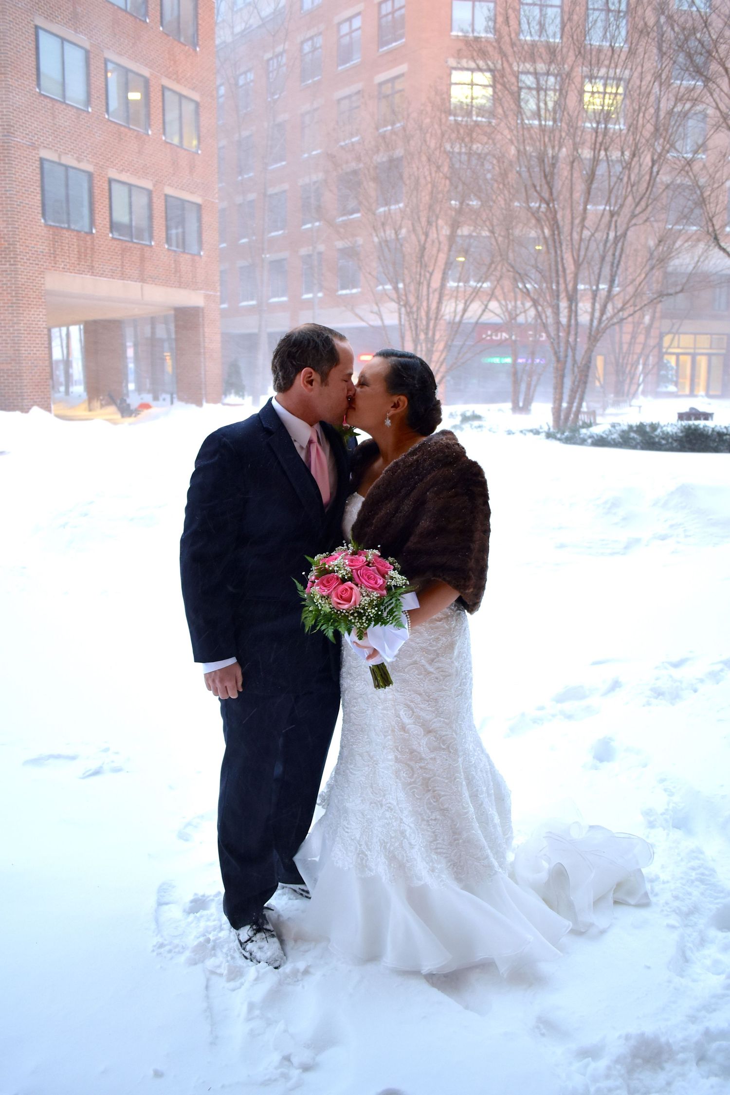 Newlyweds sharing a romantic kiss in the snow with bride holding pink bouquet.