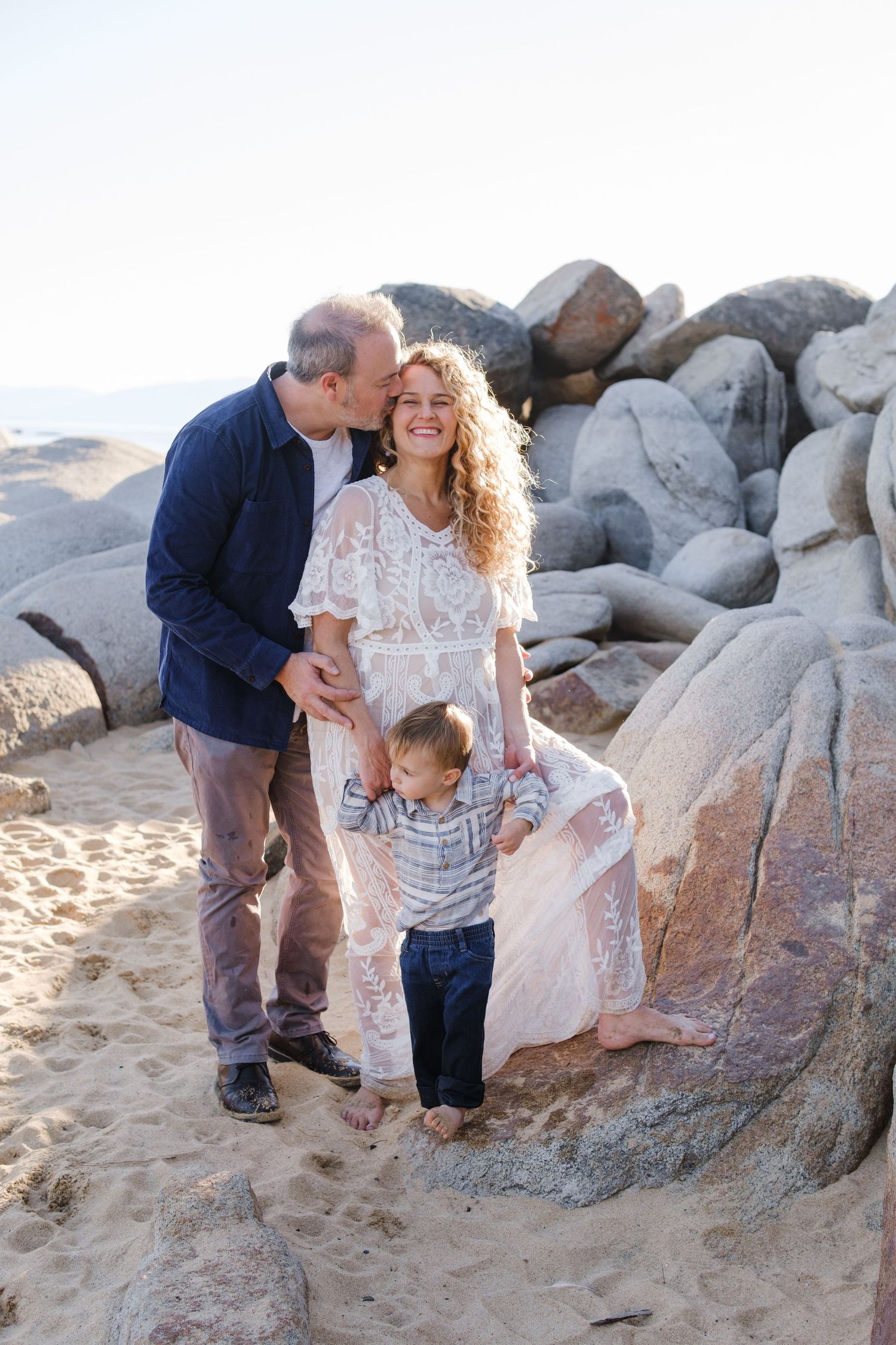 A family enjoys a tender moment together on a sandy beach in Lake Tahoe with large boulders at sunset.