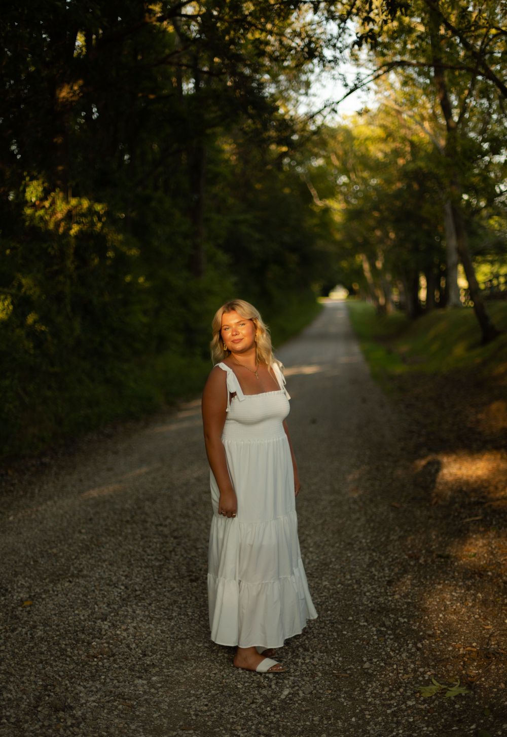 Woman in flowing white dress standing on a tree-lined country path at sunset.