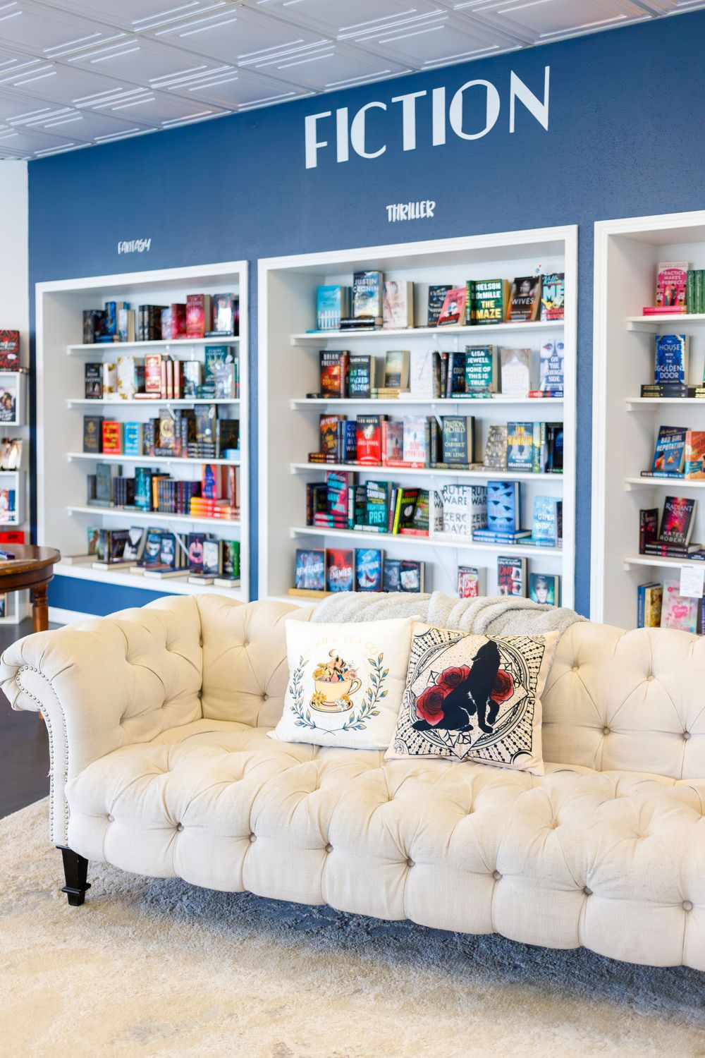 Cozy reading nook with plush tufted white sofa against blue fiction bookshelf wall in bookstore.