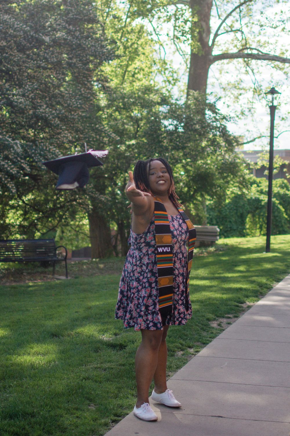 Graduate tosses cap while celebrating in floral dress on campus path.